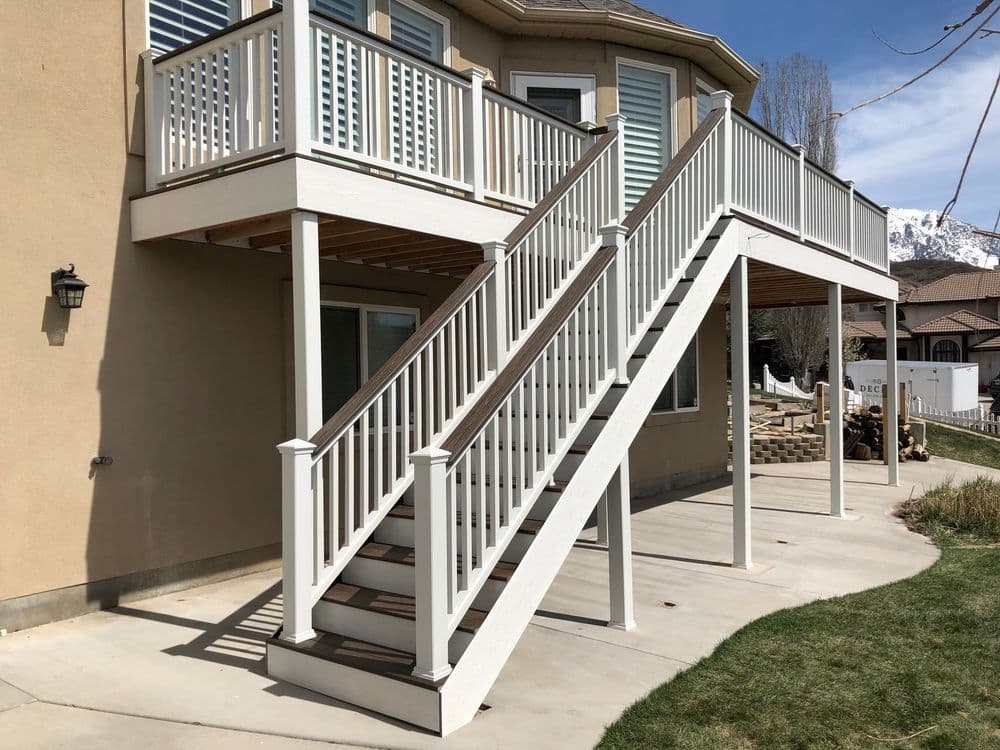 White outdoor staircase with railing leading to a second-story deck, surrounded by grass.