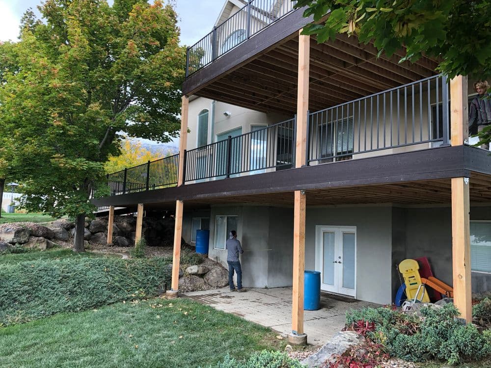 Exterior view of a two-story house with a wooden deck, greenery, and a person working below.