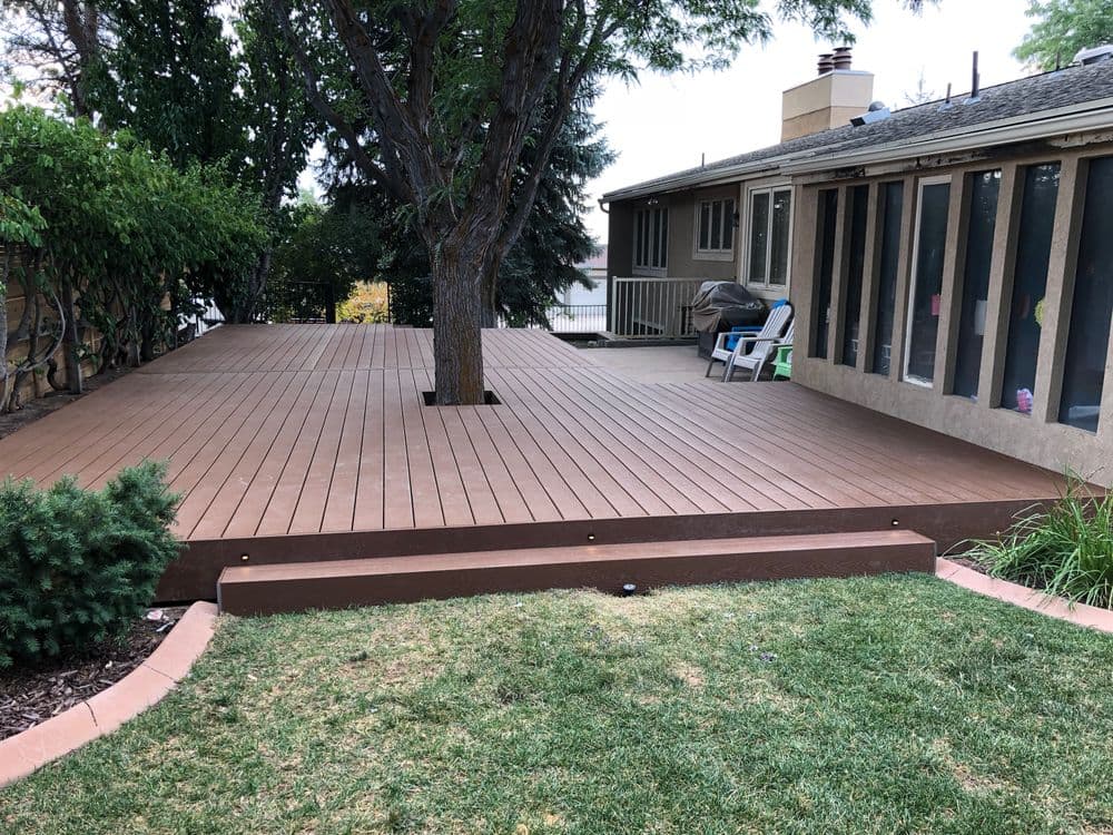 New wooden deck with tree shading, adjacent to a house, surrounded by greenery.