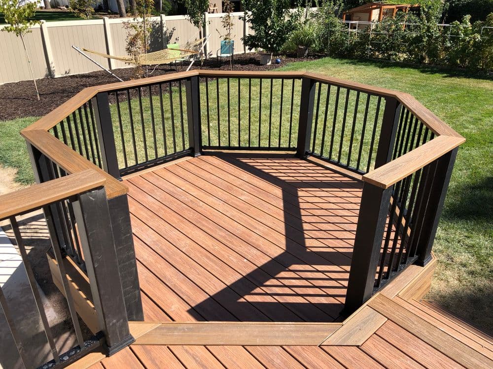 Octagonal wooden deck with black railing in a backyard setting, featuring green grass and trees.