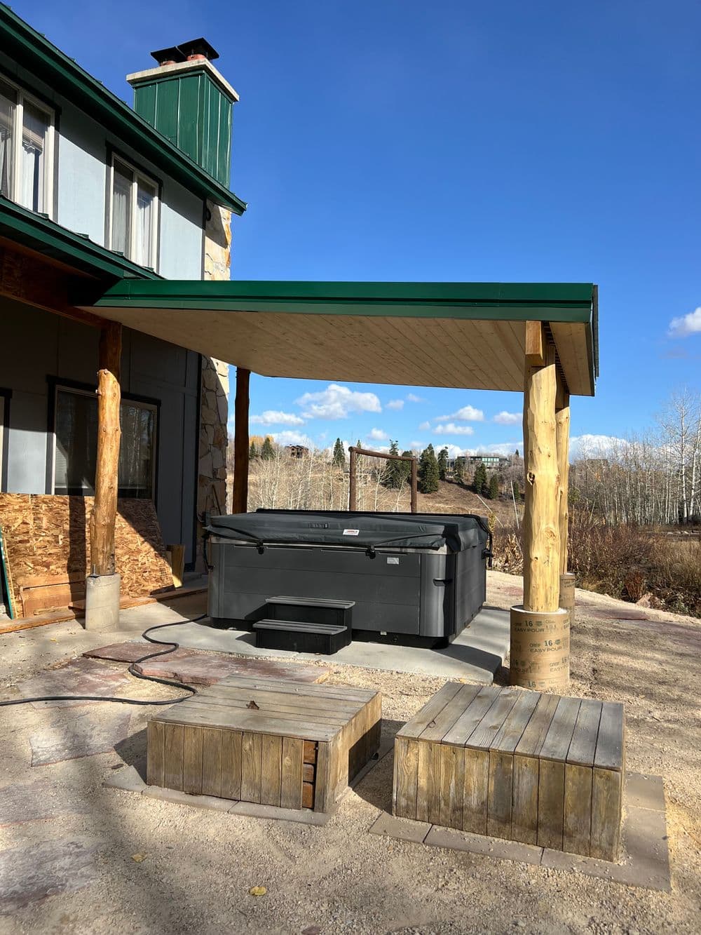 Hot tub under a wooden canopy with stone pillars, surrounded by trees and blue sky.